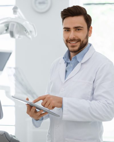 Attractive male dentist cheerful young bearded man with newest digital tablet smiling at camera, doctor using modern technologies while planning treatment, online appointment with dental clinic